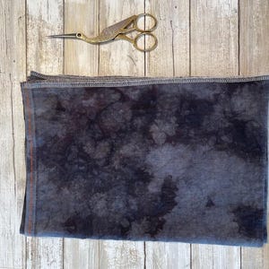 May include: A folded, dark blue fabric with a mottled pattern, possibly tie-dye, is displayed on a light wood surface. A pair of gold-colored stork scissors rests above the fabric. The fabric has a visible seam.