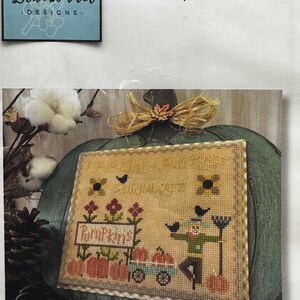 May include: Cross stitch pattern featuring a scarecrow, pumpkins, and a wagon with the text "Pumpkin Patch". The pattern is stitched on a tan fabric with a brown border.
