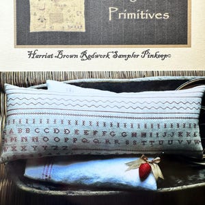 May include: A white pillow with redwork embroidery of the alphabet and numbers. The pillow is sitting on a wooden tray with a white cloth and a red strawberry. The text on the pillow reads "Harriat Brown Redwork Sampler Pinkeep".