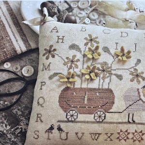 Puede incluir: Un cojín bordado a punto de cruz con una oveja tirando de un carro lleno de flores. El cojín tiene un borde alfabético y elementos decorativos. El diseño es de Brenda Gervais.
