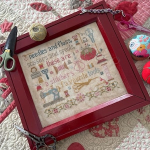 May include: A framed cross-stitch sampler with a red wooden frame. The sampler features a design of sewing tools and the text "needles and floss, scissors and spools, these are a stitcher's favourite tools".