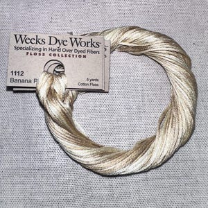 May include: A skein of light beige cotton floss, labeled "1112 Banana P" from Weeks Dye Works. The card indicates 5 yards of cotton floss. The floss is wound in a circular shape on a neutral linen background.