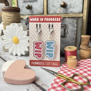 May include: Two pink and blue "WIP" (Work in Progress) stitch markers with silver lobster clasps on a white card with pink hearts and the text "Work in Progress" and "Primrose Cottage".
