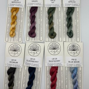 May include: Eight skeins of stranded cotton embroidery floss in various colors, including marigold, grape, ivy, spruce, aeonium, elderberry, raspberry, and blue moon. Each skein is labeled with the color name and a code. The skeins are 100% cotton and hand-dyed in Australia.