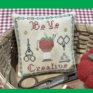 May include: A cross-stitched pillow with the words "Be Ye Creative" and needlework-themed images. The pillow is in a wicker basket with scissors and a red pincushion. The text "Antique Needleworkers" is also visible.