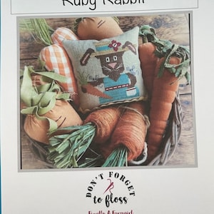 May include: Cross stitch pattern featuring a cute rabbit holding a carrot, sitting in a basket with carrots and greenery. The pattern is titled "Ruby Rabbit" and includes the text "Don't Forget to Floss" with the logo "Finally A Farmgirl".