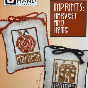 May include: Two cross-stitch pillow ornaments with brown trim. The first pillow features a pumpkin with the word "Harvest" stitched below. The second pillow features a house with the word "Home" stitched below. The pillows are part of the "Imprints: Harvest and Home" collection by Heart in Hand Needleart.