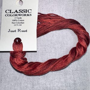 May include: A skein of Classic Colorworks embroidery floss in the color "Just Rust." The label indicates the floss is 100% cotton and 5 yards in length. The rust-colored thread is twisted into a loop, resting on a textured, off-white fabric.