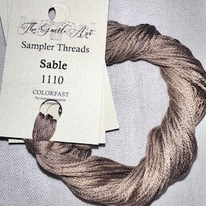 May include: A skein of Sable-colored sampler thread, with a card from "The Gentle Art" featuring the text "Sampler Threads" and "Sable 1110 COLORFAST". The thread is a mix of light and dark brown tones, and is displayed on a white fabric background.