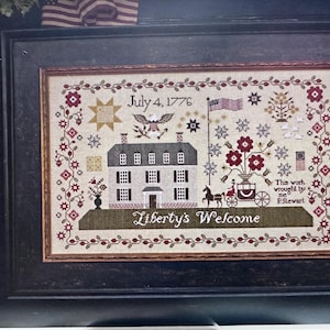 May include: Framed cross-stitch sampler titled "Liberty's Welcome" by Plum Street Samplers. The design features a colonial house, American flag, and floral elements, with the text "July 4, 1776". The sampler is in a black frame.