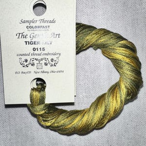 May include: A skein of "Sampler Threads" embroidery floss in shades of green and gold, labeled "TIGER LILY 0115." The floss is twisted and coiled, with a tag that reads "The Gembic Art" and "counted thread embroidery."