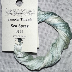 May include: A skein of "Sea Spray" sampler threads, color 0111, by The Gentle Art. The thread is a blend of pale blue, green, and white, and is wound into a circular shape. The label is off-white with black text.
