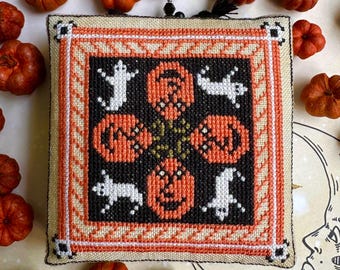 May include: A square, cross-stitched Halloween decoration featuring orange pumpkins and white ghosts on a black background. The design is framed with orange, white, and beige borders. Small pumpkins surround the decoration.