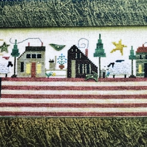 May include: A cross-stitch design featuring a village scene with houses, sheep, and trees atop a red and white striped pattern. The design includes a star, moon, and bird, with a framed border.