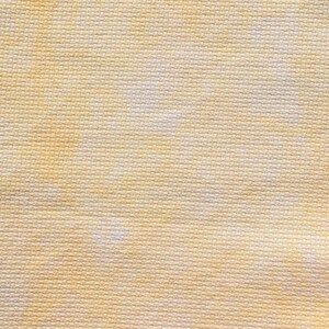May include: Close-up of a textured fabric with a woven pattern. The fabric has a light yellow color with subtle variations in tone, creating a soft, mottled appearance. The weave is tight and consistent, suggesting a durable material.