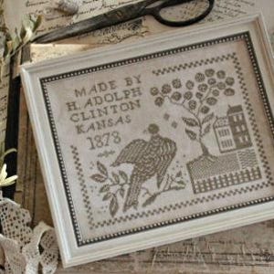 May include: Framed cross-stitch sampler with a cream-colored frame. The sampler features a bird, tree, and building design in shades of brown and tan. Text reads "MADE BY H. ADOLPH CLINTON KANSAS 1878". The artwork is displayed on a vintage background.