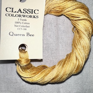 May include: A skein of golden yellow embroidery floss with a cream-colored tag. The tag reads "CLASSIC COLORWORKS" and "Queen Bee." The floss is 100% cotton and 5 yards in length. Ideal for cross-stitch and embroidery projects.