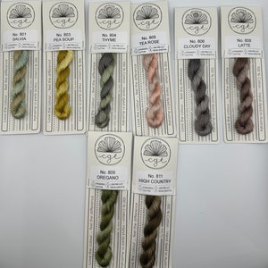 May include: A collection of 10 skeins of hand-dyed cotton embroidery floss in various shades of green, brown, yellow, and pink. Each skein is labeled with a number and a name, such as "Salvia", "Pea Soup", "Thyme", "Tea Rose", "Cloudy Day", "Latte", "Oregano", and "High Country".