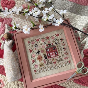 May include: A framed cross-stitch embroidery featuring a red house with a white picket fence and a garden with flowers and birds. The frame is a light pink color and the embroidery is on a cream-colored fabric.