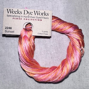 May include: A skein of Weeks Dye Works Sunset cotton floss. The floss is a blend of pink, orange, and yellow hues. The label reads "Weeks Dye Works" and "Sunset 2246". The floss is 5 yards in length.