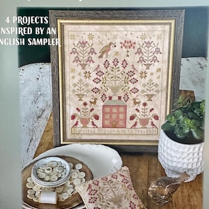 May include: A framed sampler with a detailed cross-stitch design featuring birds, flowers, and a house. The image is from "Gifts of the Season" by Blackbird Designs. Additional items include a pincushion, buttons, and a small succulent plant in a white pot.