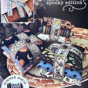 May include: A stack of decorative fabric bundles, each wrapped with embroidered ribbon and secured with a button. The bundles feature various Halloween-themed patterns, including pumpkins, moons, and stars. The text "That's A Wrap spooky edition" is visible.