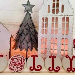 May include: A festive Christmas display featuring a metal Christmas tree, white houses with cut-out windows, and a Santa Claus figurine. The word "JOLLY" is spelled out with embroidered letters and a Santa face.