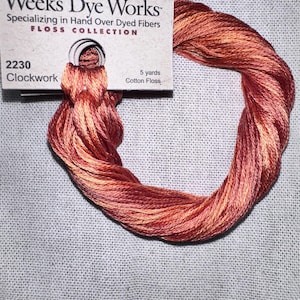 May include: A skein of Weeks Dye Works "Clockwork" cotton floss, in shades of red, orange, and yellow. The label reads "Specializing in Hand Over Dyed Fibers" and "5 yards". The floss is wound into a loop.