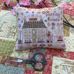 May include: A white cross-stitch pillow with a brown and green potting house design. The pillow is surrounded by pink flowers and a pair of antique scissors.