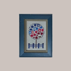 Può includere: Opera d'arte a punto croce incorniciata, con cornice blu, raffigurante un albero con una corona di stelle e forme rosse e blu. Una recinzione blu è alla base dell'albero. L'opera è su uno sfondo beige chiaro.
