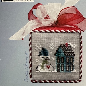 May include: A cross-stitch pattern featuring a snowman and a house in a snowy scene. The pattern is framed in a red and white border and has the text "FROSTY frill" below it.