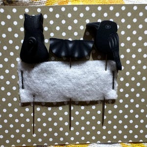 May include: Three black pincushions in the shape of a cat, a bat, and a crow. The pincushions are attached to a white felt background with black pins. The background is on a brown and white polka dot surface.