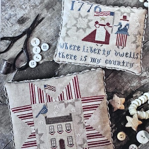 May include: Two decorative, rectangular pillows with patriotic themes. One features the text "Where Liberty Dwells" and "1776" with figures holding flags. The other has a star design with a house and flag. Additional items include scissors, buttons, and star-shaped ornaments.