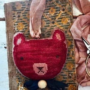 May include: A red felt bear head with a pink nose and a black bow tie. The bear is attached to a pink ribbon and is hanging on an old book.