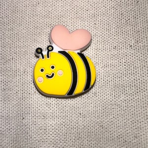 May include: A small, flat, rubberized charm shaped like a cartoon bee. The bee has a bright yellow body with black stripes, a smiling face with pink cheeks, two black antennae, and a large, light pink heart-shaped wing on its back.