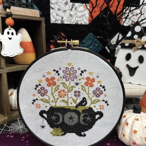 May include: A cross-stitch embroidery in a black hoop features a Halloween cauldron design. The cauldron is black with star details, and the flowers are orange, purple, and yellow. The text "Halloween Cauldron" is at the top.