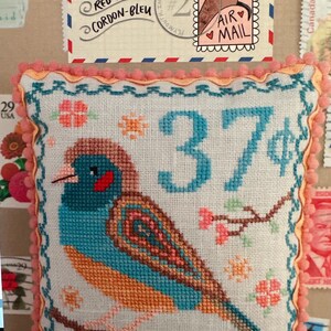 May include: A cross-stitch pillow featuring a blue and orange bird with a red cheek, perched on a branch with pink flowers. The pillow is surrounded by a pink and white trim and has the number "37" stitched in blue thread. The text "MAY RED-CHEEKED CORDON-BLEU" is visible on a stamp in the background.