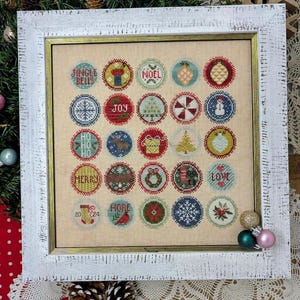 May include: A white framed cross-stitch sampler with a Christmas theme. The sampler features 24 circular designs with various Christmas motifs, including snowflakes, ornaments, reindeer, and the words "Jingle Bells", "Noel", "Joy", "Ho Ho Ho", "Merry", "Love", "20 24", and "More".