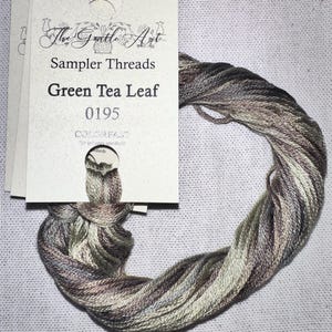May include: A skein of variegated embroidery thread in shades of green, brown, and gray, labeled "Green Tea Leaf 0195" from "The Gentle Art Sampler Threads." The thread is wound into a loose coil, with a paper tag attached.