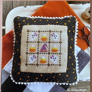 May include: A cross-stitch pillow with a Halloween theme. The pillow is made with a beige fabric and features a nine-patch design with ghosts, candy corn, and witch hats. The pillow is surrounded by a black fabric with gold stars and a white pom-pom trim. The text "October Nine Patch" is at the top of the image and "Primrose Cottage Stitches" is at the bottom.