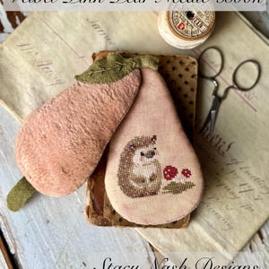May include: A needle book with a pink pear design and a hedgehog with red mushrooms. The book is on top of a vintage book with a spool of thread and scissors. The text "Velvet Pink Pear Needle Book" and "Stacy Nash Designs" are visible.