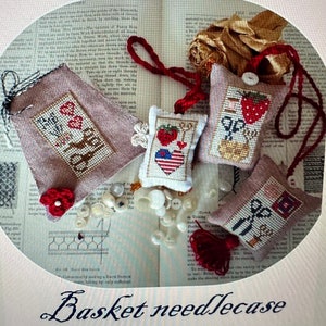 May include: A set of five small, hand-stitched needle cases with various designs. The cases are made of linen fabric and feature a variety of colors, including red, white, and blue. The cases are decorated with embroidered images of hearts, strawberries, scissors, and a teacup. The cases are attached to a red cord and are displayed on a book with white buttons scattered around them. The text "Basket needlecase" is written in black cursive at the bottom of the image.