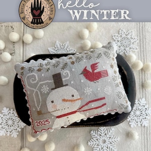 May include: A cross-stitch pillow with a snowman and cardinal design. The pillow is gray with red and white accents, and the text "2025" is visible. The words "hello WINTER" are written above the pillow.