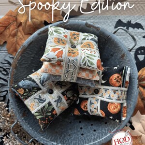 May include: A stack of decorative, square fabric items with button closures, featuring Halloween-themed patterns of pumpkins, bats, and floral designs. The items are arranged in a metal colander with fall leaves and the text "THAT'S A WRAP Spooky Edition."