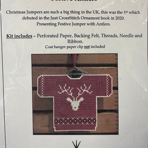 May include: A cross stitch kit for a festive Christmas jumper featuring a red sweater with white reindeer antlers and a white reindeer head. The kit includes perforated paper, backing felt, threads, a needle, and ribbon. The text on the packaging reads "Christmas Jumper Series 'Festive Antlers'".