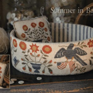 May include: A decorative, oval-shaped fabric container with a blue and white striped interior. The exterior features embroidered floral and bird designs in red, orange, and blue. The text "Summer in Baltimore" is visible.