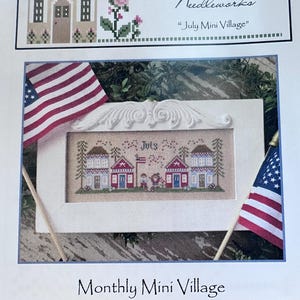 May include: A cross-stitch pattern titled "July Mini Village" featuring two houses, an American flag, and the word "July." The design is framed in white, with two small American flags on either side. The text "Country Cottage Needleworks" is at the top.