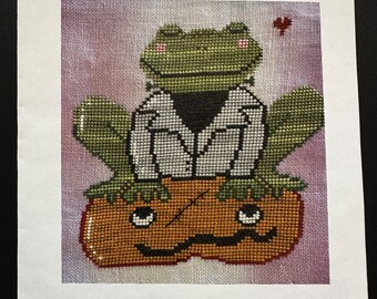 May include: Cross stitch pattern featuring a green frog wearing a white lab coat sitting on top of a jack-o-lantern. The pattern is titled "FROGGENSTEIN" and is part of a Halloween animals collection by Romy's Creations. The design is by Romina Petrucci and includes her email address.