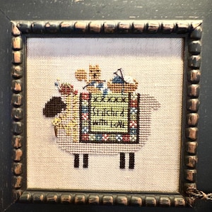 May include: A framed cross-stitch design featuring a white sheep carrying a variety of colorful items, including a small bear, a bird, and a basket. The sheep is stitched on a beige fabric and the design includes the text "Stitched with Love".