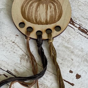 May include: A wooden pumpkin-shaped thread holder with five holes for holding embroidery floss. Three strands of floss are threaded through the holes, one brown, one black, and one gold.